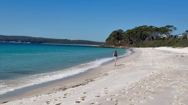 A relaxed girl walks along the white sand of Chinamans Beach, with turquoise water, gentle waves and surrounding bush, near Wollongong, Jervis Bay, New South Wales, Australia.

