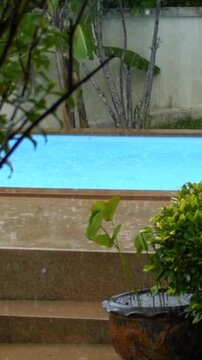 Empty swimming pool glows turquoise while rain falls past potted plants and steps in tropical garden. Quiet poolside setting feels serene, with wet stone, leafy branches, and muted wall behind.