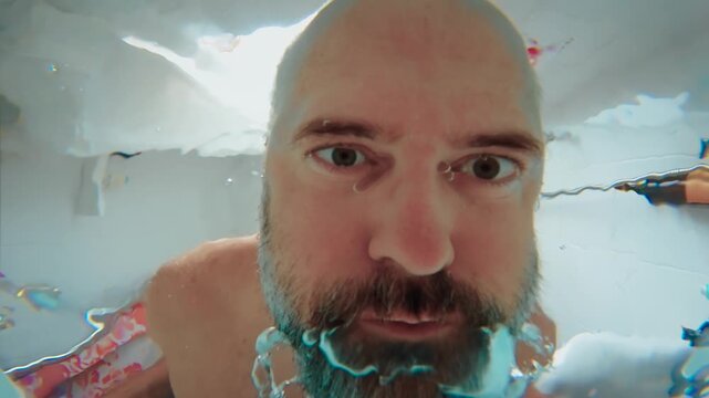 Depressed man lowering head into bathtub water, view from bottom of bathtub. Bearded man looking underwater with serious expression in bathroom. Mental health crisis concept with submerged face and