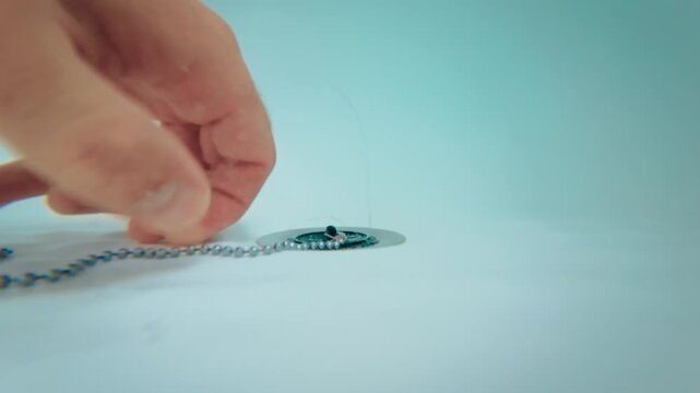 Hand opening bathtub drain by lifting plug on chain for water release, view from bottom of bathtub. Close up fingers removing rubber stopper from drain hole. Bathroom plumbing, drainage and manual