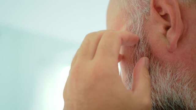 Gray beard inspection with man running fingers along facial hair near cheek and jaw. Close up male grooming routine focused on beard texture and skin. Middle aged man checking stubble, facial hair