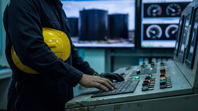 Industrial process control and plant monitoring concept, operator using a console keyboard while holding a yellow hard hat beside illuminated switches and gauge displays in a factory control room