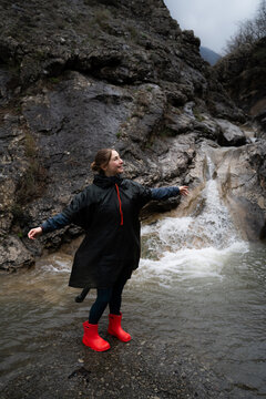 Smiling woman enjoying a walk in a rocky mountain gorge during cloudy weather, dressed in waterproof clothing and rubber boots. Outdoor recreation, adventure travel and freedom concept.