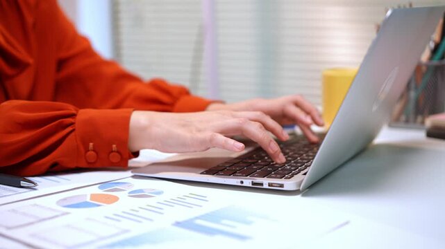 Close up of woman's hands typing on laptop in modern office with business charts and reports on desk