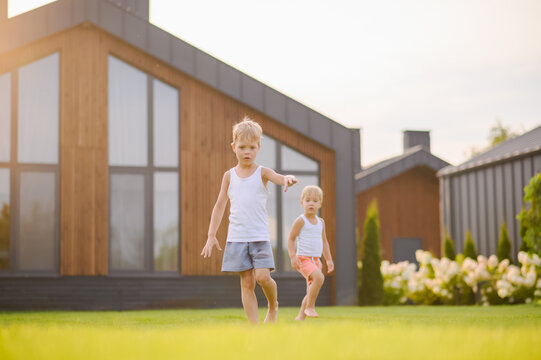 Young children running barefoot and playing together on green lawn near a modern house in warm evening sunlight. Active childhood, outdoor play, summer leisure and carefree lifestyle concept.