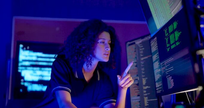 Adult female technology analyst rests hand near chin while studying multiple code monitors, then resumes work during cybersecurity monitoring, digital system analysis, and secure business data review.