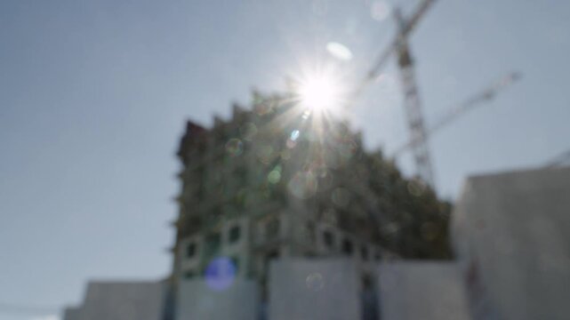 Mid-construction concrete high-rise shell with cranes backlit by sun, exposed floor slabs and column grid, temporary formwork at upper levels, lens flare and starburst