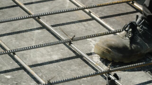Low-angle close up of gloved hand preparing tie wire coil and worn work boot on reinforced rebar grid over concrete foundation slab in bright sunlight outdoors in daylight
