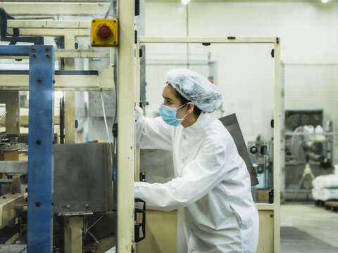 Female Factory Operator Inspecting Packaging Machine Food Process Plant Female technician in protective hygienic coat hairnet mask operating industrial packaging machinery quality assurance concept.