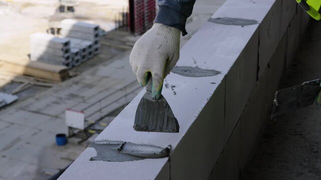 Gloved hand spreads gray mortar with notched trowel across top of white aerated concrete block wall at outdoor construction site