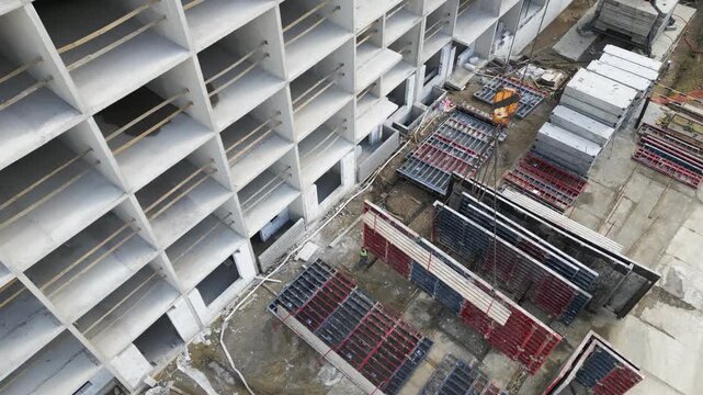 Aerial oblique view of crane lifting stacked formwork panels beside multistory precast concrete building at construction site