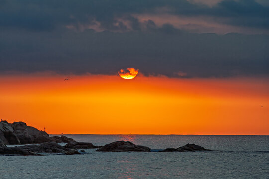 Dramatic sunrise with a fiery orange sky and half sun peeking under heavy dark clouds over the rocky coast of Platja d'Aro, Costa Brava, Spain.