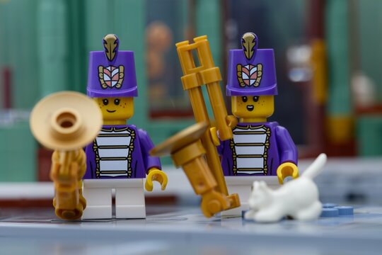 Tambov, Russian Federation - July 14, 2026 Two Lego marching band musician minifigures wearing purple uniforms and tall hats hold golden wind instruments  and white cat on a city street.