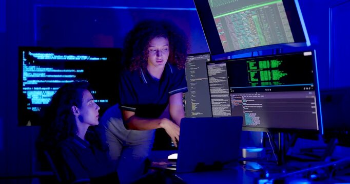 Adult female technology colleagues review source code on monitors, one leaning toward seated coworker while both discuss cybersecurity analysis and system monitoring in digital operations office.