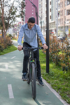 A young man is standing next to his bike on a pathway in a city. He wears a light blue shirt and black pants.