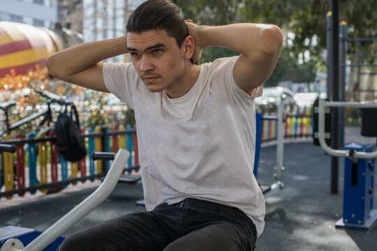 A young man does a workout routine using exercise equipment at an outdoor gym in a park during daylight.