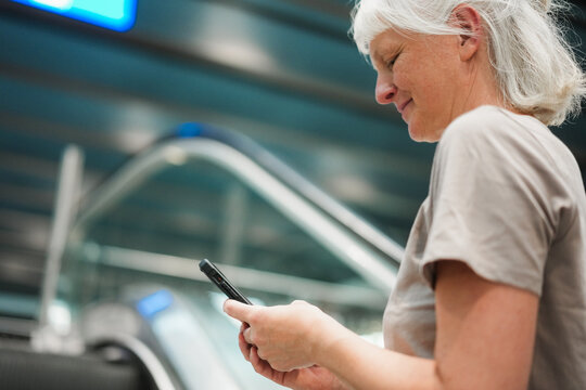 Caucasian 50's woman using smartphone on escalator inside train station wearing casual clothes - City lifestyle and technology concept - Main focus on female ear