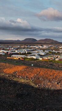 Vertical drone view of Grindavik, Iceland, showing a protective lava berm between dark volcanic terrain and a residential district under dramatic clouds and warm evening light.