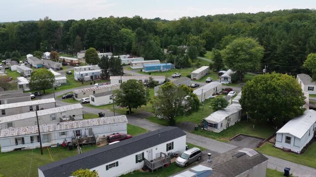 A mobile home park meets thick summer woodland on the edge of Lynchburg, Virginia, its rows of pale trailers giving way to an unbroken green tree line under soft cloud. Aerial rising wide shot
