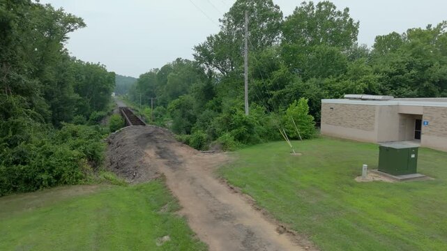A view shows a railway track stretching through a green area. Located near a building, dirt and trees are seen on either side. The setting captures nature and infrastructure in a rural landscape.