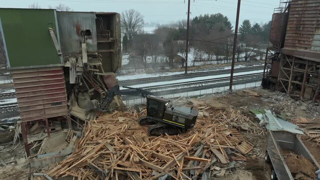 A machine removes wooden debris at a demolition site near train tracks. The area is busy as workers manage the cleanup task. Smoke and dust fill the air during the process.