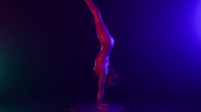 Female acrobat in a shiny bodysuit performs a dramatic handstand split on a pedestal, illuminated by vibrant blue and magenta studio lights on a dark stage.