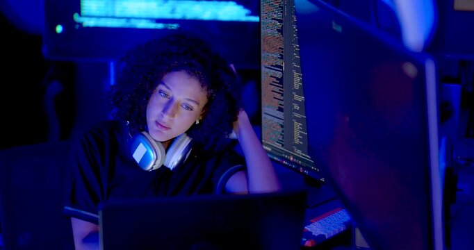 Adult female technology professional with headset around neck studies laptop and code monitors, touches hair and resumes screen review during cybersecurity monitoring and digital system analysis.