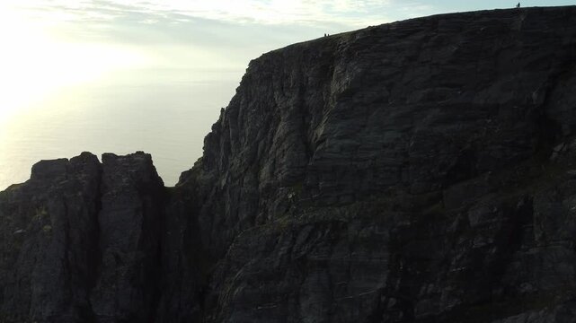 Aerial ascent past the towering silhouetted cliffs of Nordkapp in Norway, revealing tiny tourist figures standing at the cliff edge against a bright sunlit sky over the sea