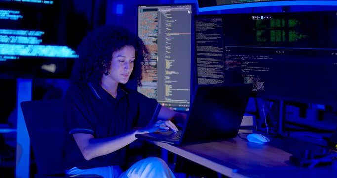 Adult female technology professional types on laptop, smiles briefly, reviews source code across monitors and touches forehead while continuing cybersecurity monitoring in digital operations office.