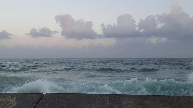 Foaming sea waves break and splash over rocky shallows near the concrete seawall of historic Jaffa Port along the Mediterranean coast under an overcast daytime sky Pan Shot