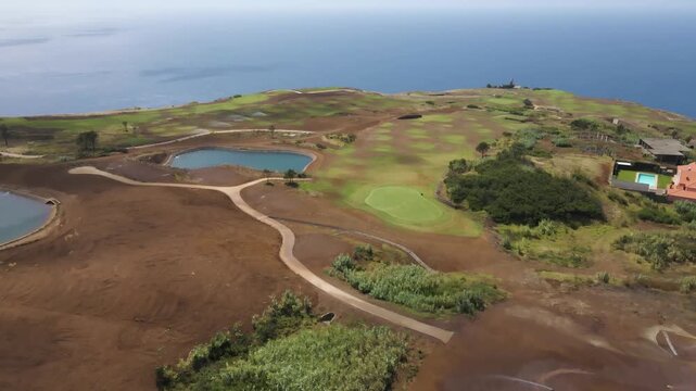 Top-down drone shot capturing geometric patterns of greens and bunkers contrasting with wild coastal cliffs and deep blue sea. Drone moving backwards over the golf course showing some green patches.