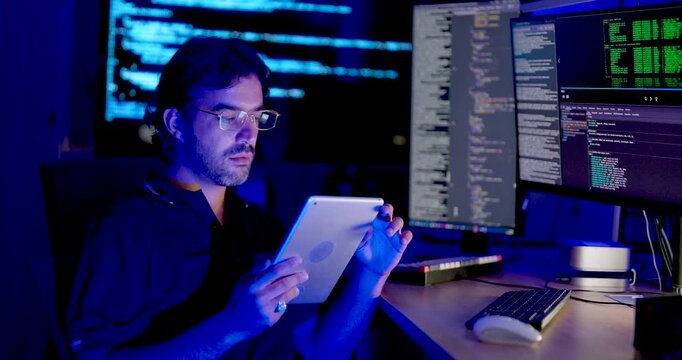 Adult Asian male cybersecurity analyst holds tablet while using mouse and checking multiple code monitors, combining mobile data review with digital system monitoring and network security analysis.