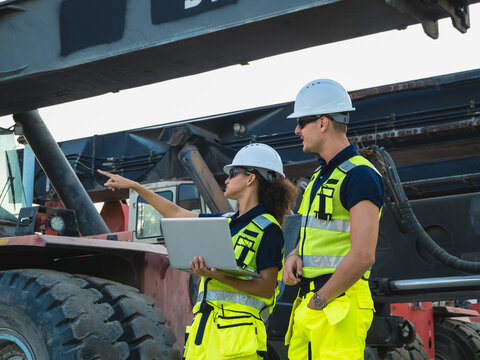 Diverse Logistic Engineer with Laptop Inspecting Container Reach Stacker: Male and female port supervisor in hardhat and hi-vis gear directing cargo freight operation supply chain concept.