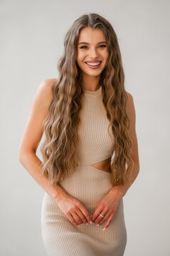 Portrait of smiling young woman with long wavy hair wearing beige ribbed dress posed against neutral studio backdrop, showcasing natural makeup, friendly expression, and confident relaxed posture