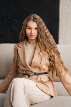 Elegant woman seated on sofa wearing beige coat over ribbed knit dress, calm fashionable portrait with warm soft lighting against dark studio background