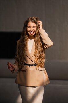 Smiling woman in beige blazer and knit dress outdoors in warm sunlight, professional fashion portrait with soft shadows, confident pose, neutral street background