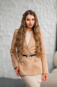 Portrait of stylish woman in beige trench coat standing outdoors against grey textured wall, showcasing relaxed confidence, soft makeup, long wavy hair, fashion-forward look, elegant neutral ensemble