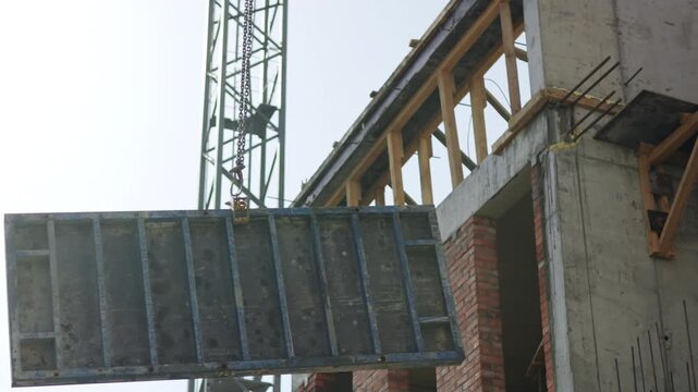 Tower crane lifts heavy concrete block near building carcass on gloomy day. Machine transports cargo as works at urban construction site