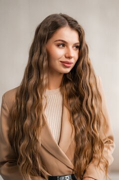 Portrait of woman with long wavy brown hair wearing beige blazer and belt, looking away against neutral backdrop, soft lighting, natural makeup, fashion portrait with elegant styling
