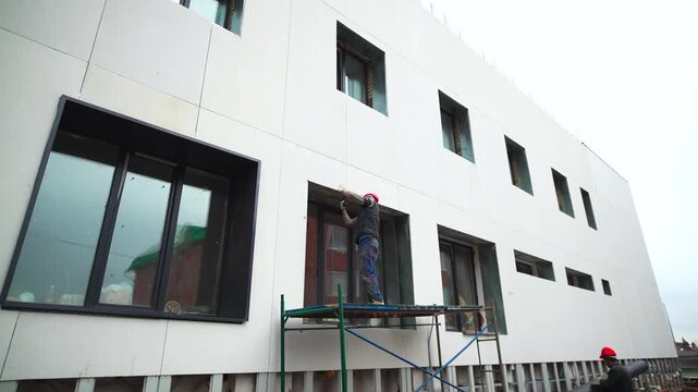 Builders work with commercial building facade at construction site slow motion. Laborers install decorative materials on window frame