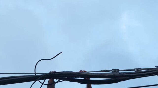 Pan right video of an electrical worker hands holding twisted aerial cables after completing a joint, industrial power grid distribution network maintenance against a blue sky.