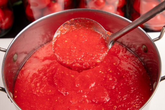 Process of making homemade tomato sauce close up. Ladle scooping hot boiling red tomato paste, lecho or adjika from metal pot. Horizontal food preserving and canning photography.