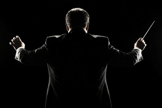 Back view of male conductor in black suit conducting orchestra with baton. Man stands in dark studio holding music wand with arms wide. Monochrome silhouette of pro musician leads classical symphony
