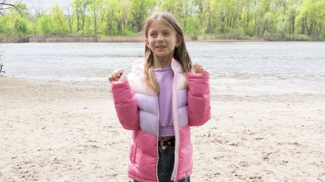 Cute Little Girl Posing on a Sandy River Beach in a Colorful Puffer Jacket