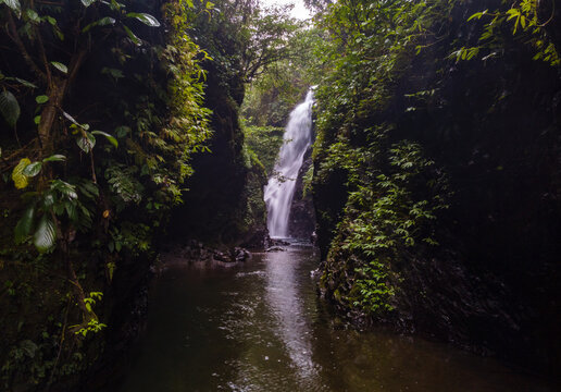 Aerial View of Hidden Lesung Waterfall Deep in the Jungle on the Slopes of Mount Slamet, A Cave-Like Tunnel Formed by Wild Trees, Moss, and Rocks Frames the Waterfall in Indonesia.