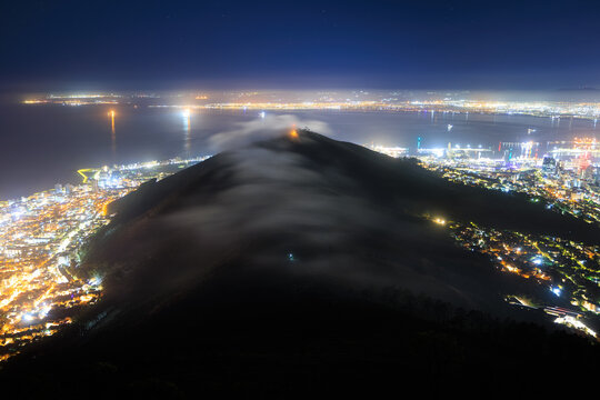 View of Signal Hill at night with clouds rolling over the ridge and city lights illuminating the coastline in Cape Town, Western Cape, South Africa.