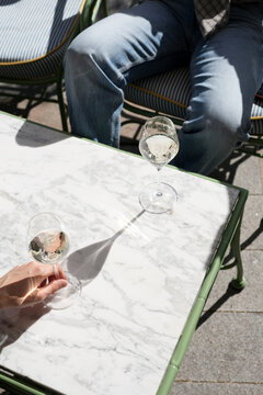 Man's hand holding a glass of chilled white wine or sparkling champagne at a marble bistro table on a sunny outdoor terrace.