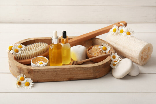 Spa composition with tray, massage body brush and bottles of chamomile essential oil on white wooden background, closeup