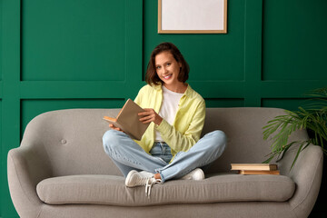 Pretty young woman reading book on sofa near green wall