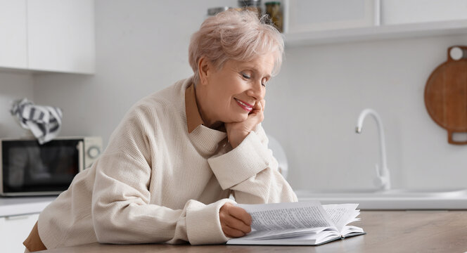 Healthy aging and retirement lifestyle, happy senior woman reading a book at home in a modern kitchen for leisure and mental wellness